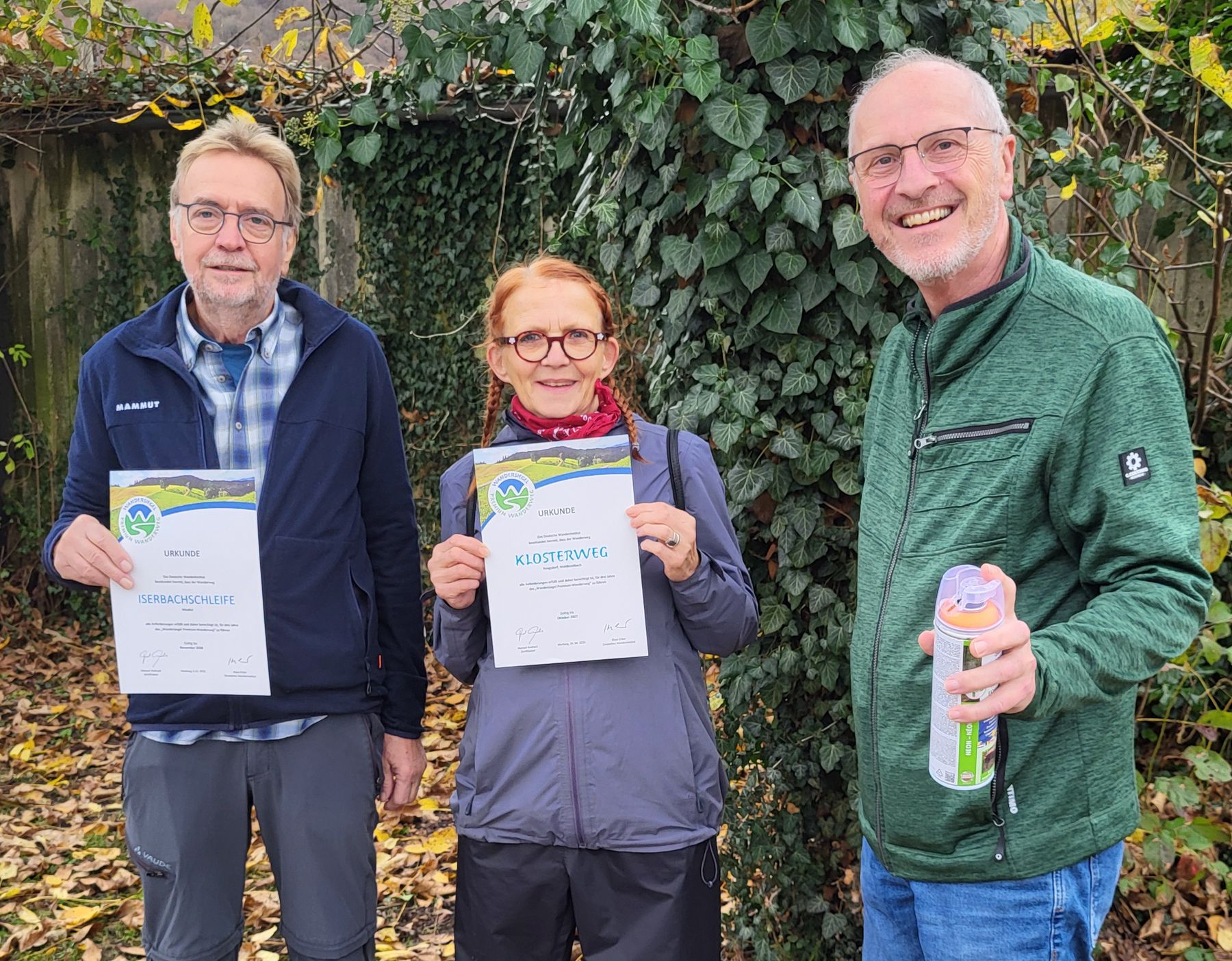 Three smiling adults stand outdoors on fallen leaves, two holding certificates and one holding a can. They are dressed in casual jackets and glasses, with greenery and ivy in the background.